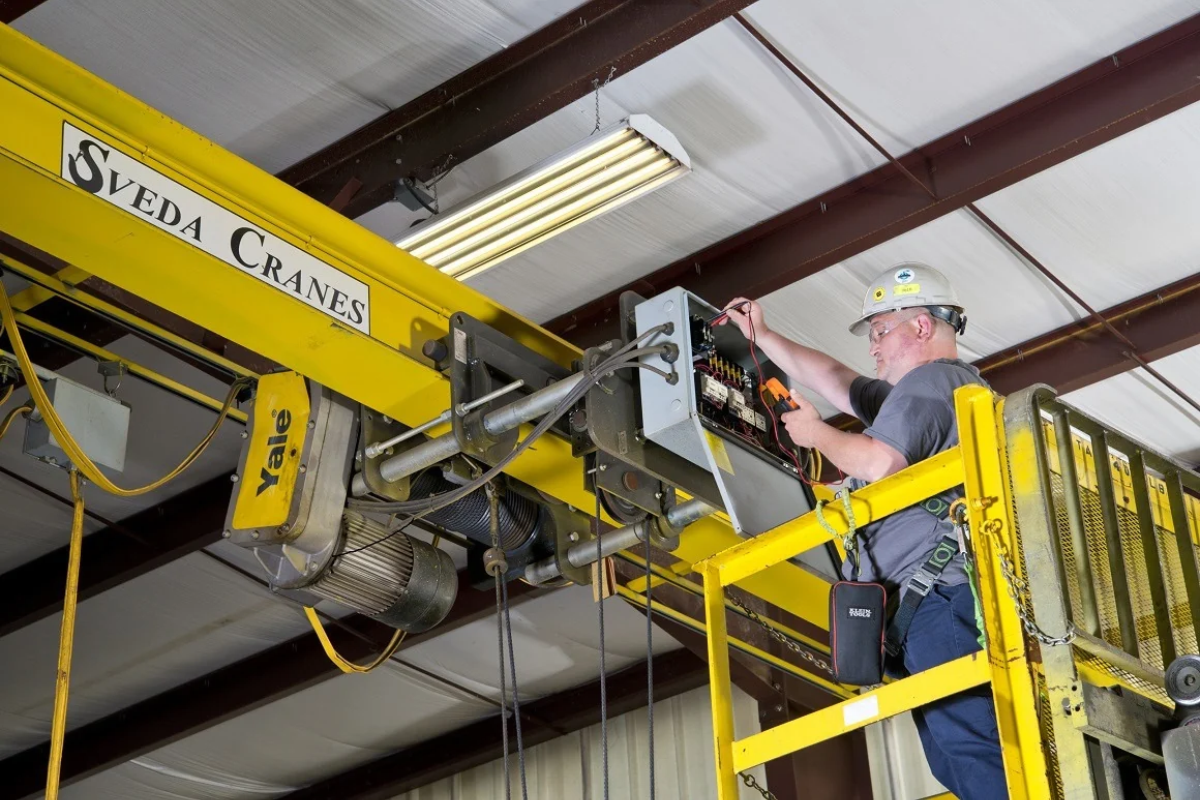 Industrial crane technician wearing safety equipment works from an elevated yellow lift to inspect and service the electrical control panel of an overhead bridge crane inside a manufacturing facility.